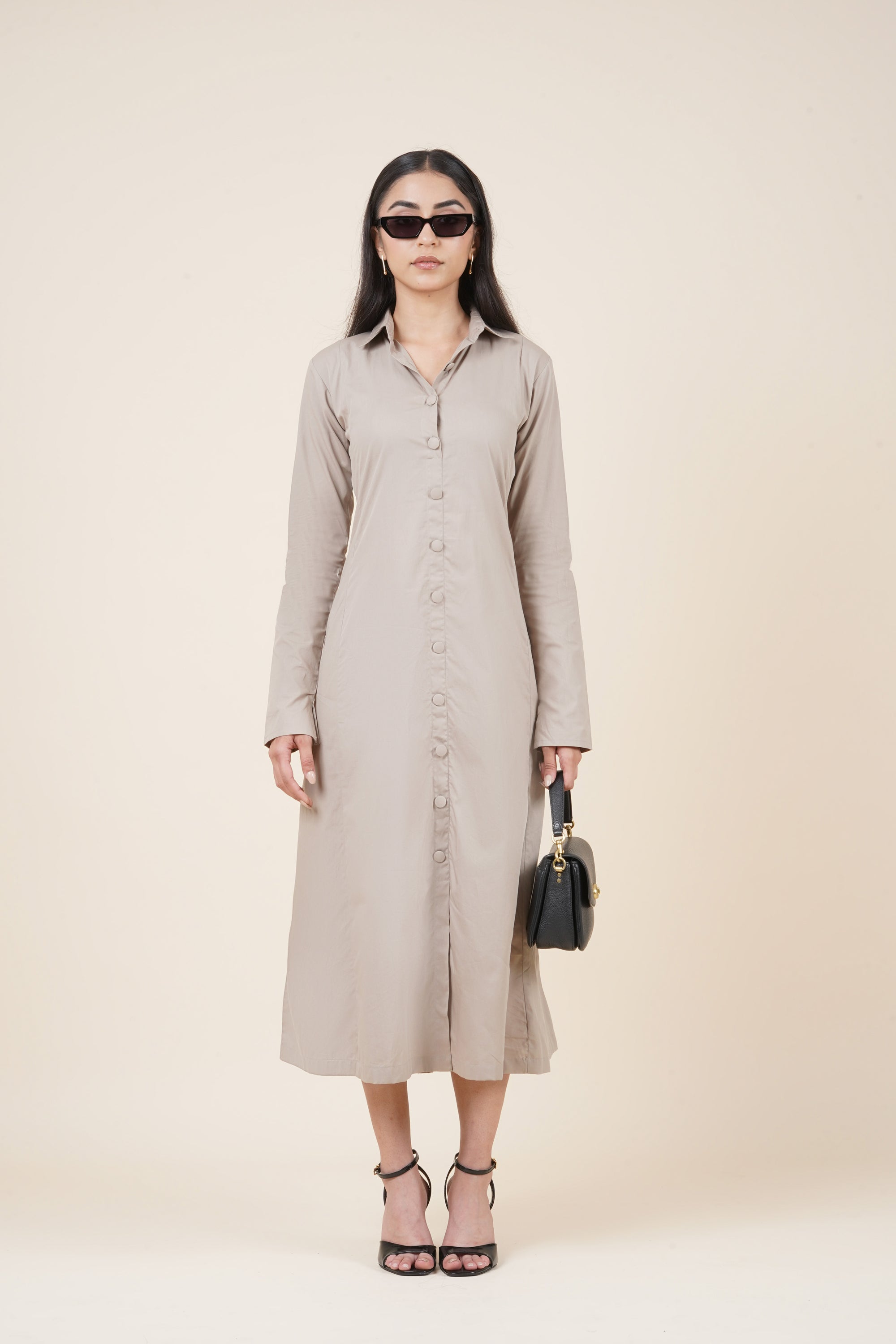 Woman wearing a beige button-up dress with sunglasses and a black handbag on a plain background
